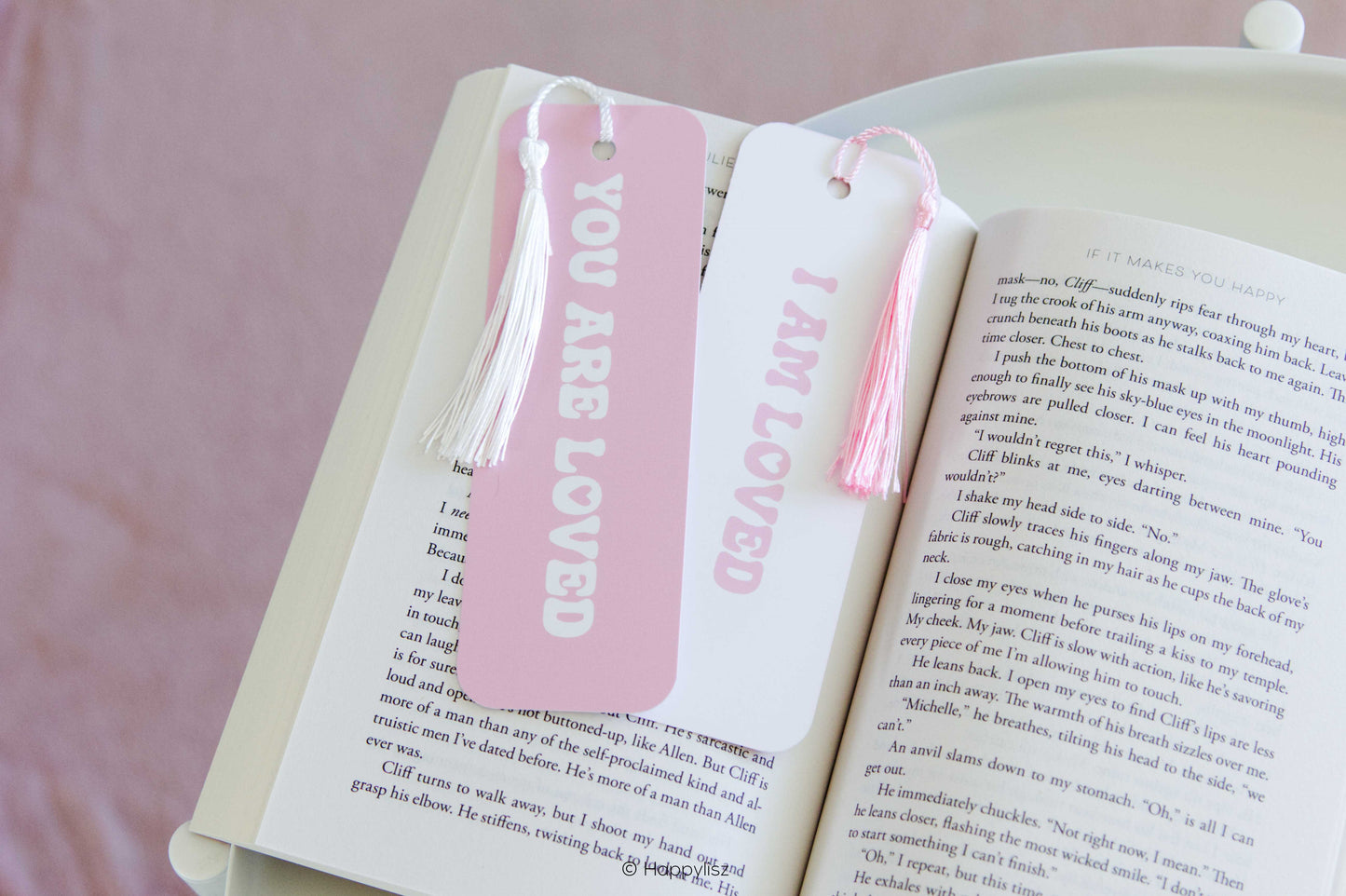Two decorative bookmarks shaped like book pages, one pink and one white, both with tassels and the words 'YOU ARE LOVED' and 'I AM LOVED' printed vertically, resting on an open book.