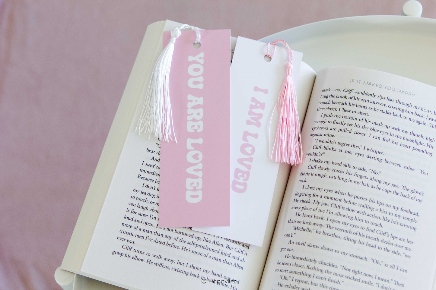 A pair of white book bookmarks with pink tassels and pink lettering, shown bookmarking an open book on a soft pink background. One bookmark reads 'YOU ARE LOVED' and the other reads 'I AM LOVED'.