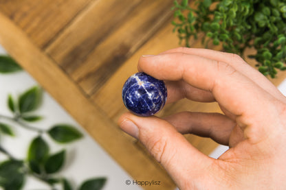 Hand holding a small blue stone with a blurred background of green plants and wooden surface.